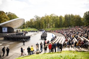amfiteatr na powietrzu. Przed scena w szeregach stoją umundurowani litewscy policjanci i zaproszeni goście. Stojąca z boku grupka ludzi i siedzący na trybunach przyglądają się policjantom