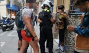 Ulica w Barcelonie. Dwaj umundurowani hiszpańscy policjanci stoją przed mężczyzną, który ma uniesioną jedną rękę do góry. Za policjantami stoją dwaj mężczyźni w krótkich spodenkach.