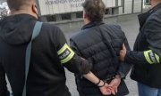 Dziedziniec komendy. Dwóch policjantów po cywilnemu z opaskami na rękawach z napisem Policja, prowadzi zatrzymanego, który ma założone na rękach trzymanych z tyłu kajdanki