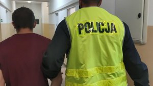 Policjant w żółtej kamizelce z czarnym napisem Policja prowadzi korytarzem zatrzymanego mężczyznę