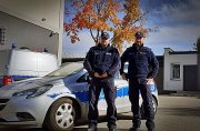 Dwaj umundurowani policjanci stoją przy radiowozie. Zdjęcie na zewnątrz. W tle budynki i drzewo z kolorowymi liśćmi