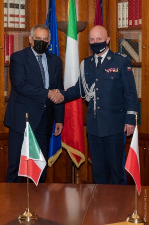 dwaj mężczyźni ściskają sobie dłonie i pozują do zdjęcia. Mężczyzna z lewej strony ubrany jest w garnitur, mężczyzna z prawej w mundur generała polskiej Policji. Za nimi widoczne są dwie stojące flagi Włoch i Unii Europejskiej, a przed nimi na stole ustawione są flagi Włoch i Polski