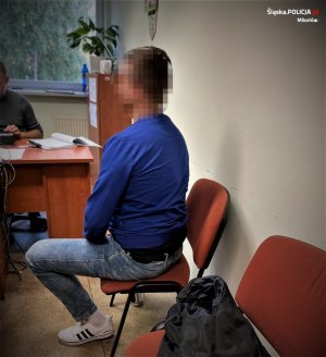 na krześle siedzi mężczyzna w niebieskim swetrze, za biurkiem siedzi policjant wydziału kryminalnego