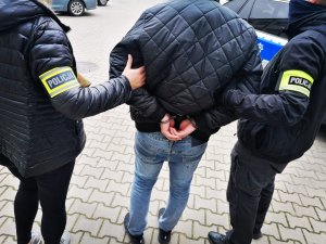 dwoje policjantów - kobieta i mężczyzna - w ubraniach cywilnych i opaska na ręce z napisem Policja, prowadzi zatrzymanego mężczyznę