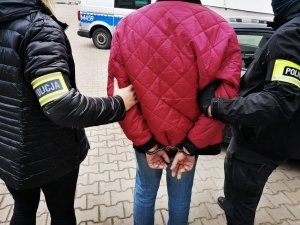 dwoje policjantów - kobieta i mężczyzna - w ubraniach cywilnych i opaska na ręce z napisem Policja, prowadzi zatrzymanego mężczyznę