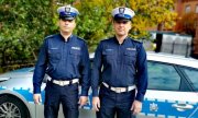 dwaj umundurowani policjanci ruchu drogowego pozują do zdjęcia, z animi stoi policyjny radiowóz