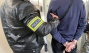 Policjant ubrany po cywilnemu z opaską z napisem Policja trzyma skutego w kajdanki mężczyznę