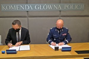 mężczyzna w garniturze i generał Policji siedzą przy stole i podpisują lezące przed nimi dokumenty. Nad mężczyznami widać napis: Komenda Główna Policji