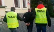 dwaj policjanci w żółtych kamizelkach z napisem Policja prowadzą zatrzymanego skutego kajdankami