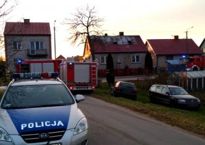 radiowóz policyjny, dwa wozy strażackie i dwa samochody osobowe przed budynkami