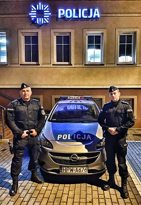 dwaj policjanci stoją przy radiowozie w tle budynek komendy policji