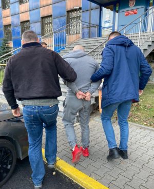 zdjęcie kolorowe: na chodniku przed wejściem do Komendy Miejskiej Policji w Katowicach policjanci wydziału kryminalnego prowadzą zatrzymanego mężczyznę poszukiwanego europejskim nakazem aresztowania, któremu założono kajdanki zespolone