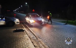 Policjant ruchu drogowego z latarką koloru czerwonego w nocy podchodzi do zatrzymanego samochodu, obok drogi stoi radiowóz policyjny