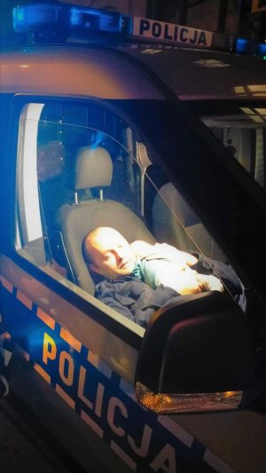 Zdjęcia zrobione na zewnątrz - ukazuje wnętrze radiowozu, w którym siedzi policjant, a na ramieniu trzyma śpiącego niemowlaka