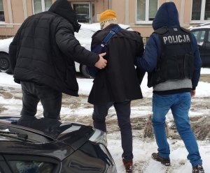 Policjanci prowadzą zatrzymanego mężczyznę. Jeden z nich ma na plecach napis Policja. Widok z tyłu
