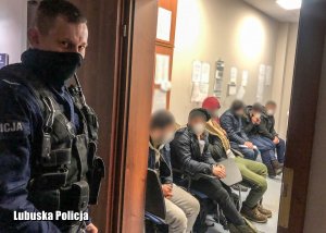 Policjant na korytarzu w komendzie, a w pomieszczeniu mężczyźni siedzący na krzesłach - twarze mężczyzn zamazane graficznie