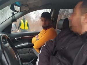 dwaj mężczyźni w samochodzie, z tyłu widać policjanta w żółtej kamizelce z napisem Policja