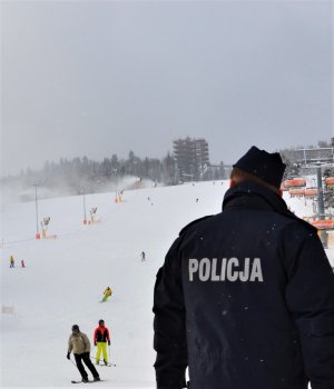 na pierwszym planie policjant prewencji odwrócony tyłem w tle narciarze zjeżdżający ze stoku