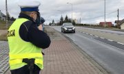 policjant ruchu drogowego trzyma w ręku ręczny miernik prędkości i mierzy prędkość jadącego po jezdni pojazdu