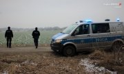 radiowóz policyjny, w tle umundurowani policjanci na polu w trakcie akcji poszukiwawczej