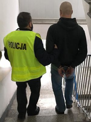 policjant w żółtej kamizelce odblaskowej z napisem policja prowadzi po klatce schodowej zatrzymanego mężczyznę