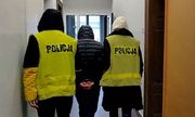 Dwoje policjantów w żółtych kamizelkach z napisem Policja na plecach, korytarzem prowadzi zatrzymanego mężczyznę