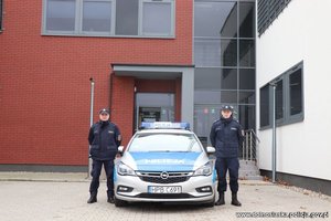 dwaj umundurowani policjanci stoją przed budynkiem komisariatu przy radiowozie