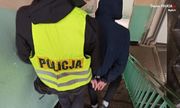 Policjant pionu kryminalnego z założoną kamizelką z napisem Policja prowadzi zatrzymanego, który ma założone kajdanki na ręce trzymane z tyłu