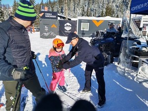 dziecko w stroju narciarskim z założonymi nartami, policjant zapinający na ręce dziecka opaskę odblaskową