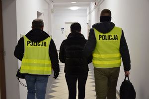 Dwaj funkcjonariusze w kamizelkach z napisem POLICJA, prowadzą przez korytarz zatrzymaną kobietę