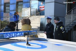umundurowany policjant i kierowca rajdowy Jarosław Szeja - stoją przed budynkiem komendy wojewódzkiej, w tle stoi mężczyzna z kamerą