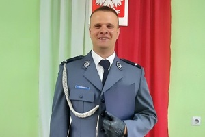policjant w galowym mundurze pozuje do zdjęcia na tle zawieszonej za nim biało czerwonej flagi i godła Polski