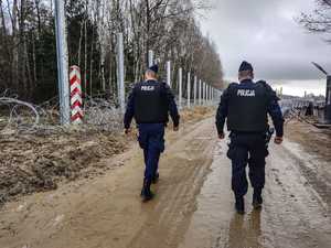 policjanci idą wzdłuż budowy zapory granicznej, po lewej stronie widoczny słupek graniczny
