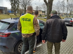 Dwaj policjanci, jeden ubrany po cywilnemu, drugi w żółtej kamizelce z napisem Policja na plecach prowadzą zatrzymanego