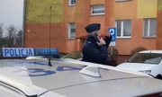 umundurowany policjant stoi przy radiowozie i trzyma telefon komórkowy przy uchu, w tle blok mieszkalny