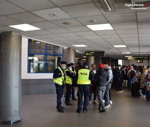 policjanci w mundurach i kamizelkach odblaskowych udzielający informacji grupie osób, w tle duża liczba osób w holu terminala lotniskowego