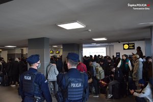 policjanci w mundurach obserwujący liczną grupę pasażerów czekających w kolejce, w terminalu lotniska