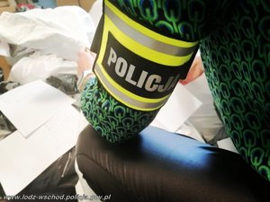 widok na opaskę z napisem policja, którą policjantka pochylona nad zabezpieczonymi ubraniami popakowanymi w worki ma na lewym przedramieniu