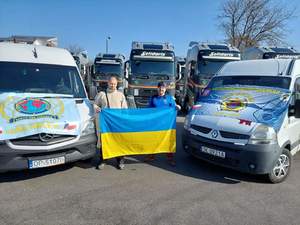 dwaj mężczyźni z flagą Ukrainy stoją pomiędzy dwoma autami, które na maskach maja rozłożone flagi IPA