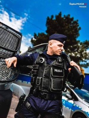 Zdjęcie kolorowe: umundurowany policjant, w tle widoczny radiowóz