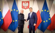 oficer Policji ściska dłoń mężczyźnie w garniturze Za nimi z obu stron widoczne są flagi unijne i Polski w stojakach