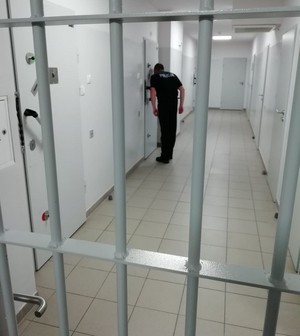 Policjant w izbie zatrzymań zagląda do celi przez wizjer. na pierwszym planie krata aresztu