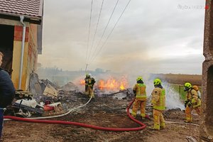 strażacy dogaszają pożar drewnianych przedmiotów, pożar wybuchł blisko ściany budynku gospodarczego, z boku stoi mężczyzna i obserwuje pracę strażaków