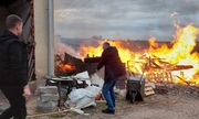 w tle pożar drewnianych przedmiotów, mężczyzna wodą z węża ogrodowego polewa ogień, drugi mężczyzna zbliża się do niego. Pożar znajduje się obok ściany budynku gospodarczego