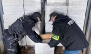policjant CBŚP i funkcjonariusz KAS otwierają karton z papierosami, w tle na paletach białe kartony