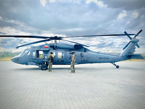 Na płycie lądowiska policyjny Black Hawk sfotografowany z lewego boku przed nim dwóch funkcjonariuszy w umundurowaniu taktycznym.