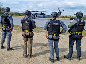 Czterech funkcjonariuszy widzianych od tyłu, którzy przyglądają się stojącemu na płycie lądowiska Black Hawkowi.