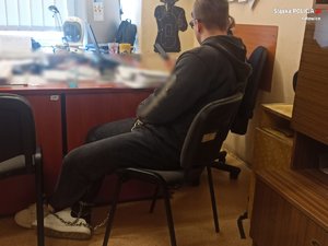 zatrzymany mężczyzna z założonymi na ręce i nogi kajdankami zespolonymi, siedzący na krześle podczas przesłuchania