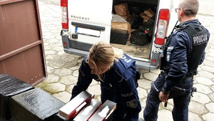 policjanci na dziedzińcu wynoszą zabezpieczone paczki nielegalnych papierosów z busa koloru białego