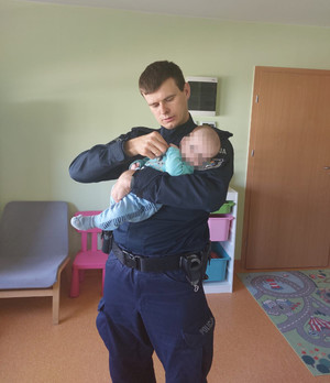 Zdjęcie przedstawia policjanta w granatowym mundurze, który trzyma w ramionach małe dziecko w niebieskich śpioszkach. Twarz dziecko jest zasłonięta komputerowym filtrem